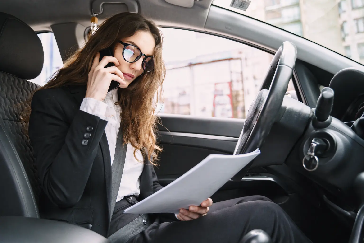 Businesswoman in glasses talking on phone while reviewing documents in car